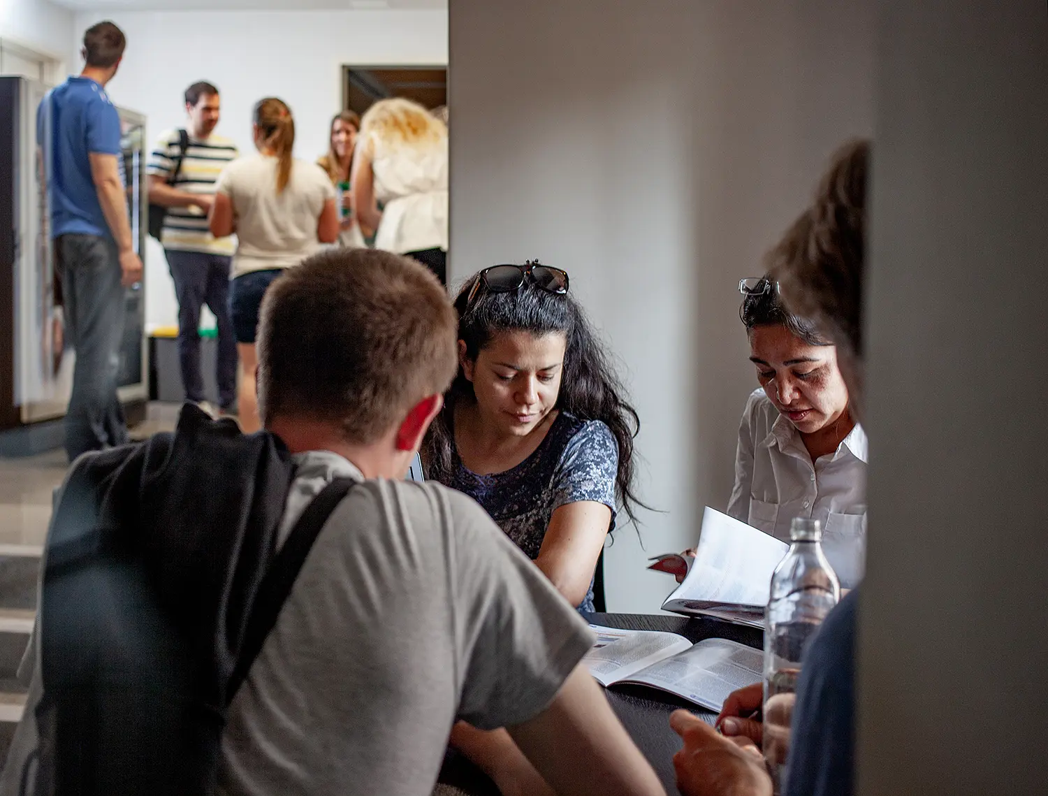 Vier Personen sitzen an einem Tisch mit Arbeitsmaterial. Andere stehen vor einem Getränkeautomaten.