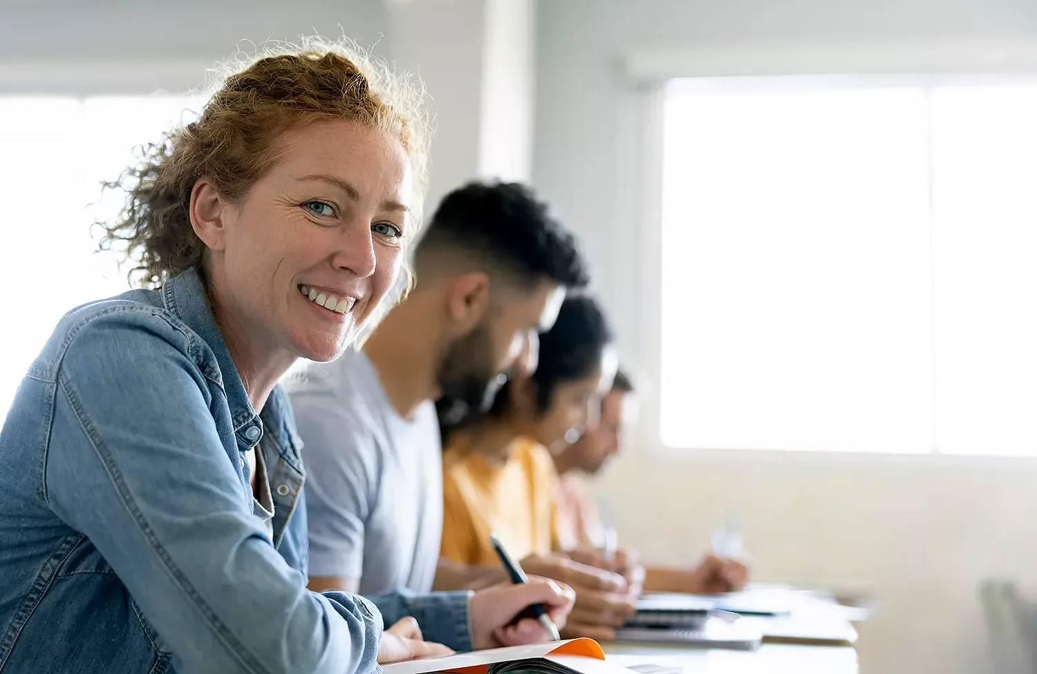 Vier Erwachsene sitzen schreibend am Tisch. Eine Frau im Vordergrund lacht in die Kamera.