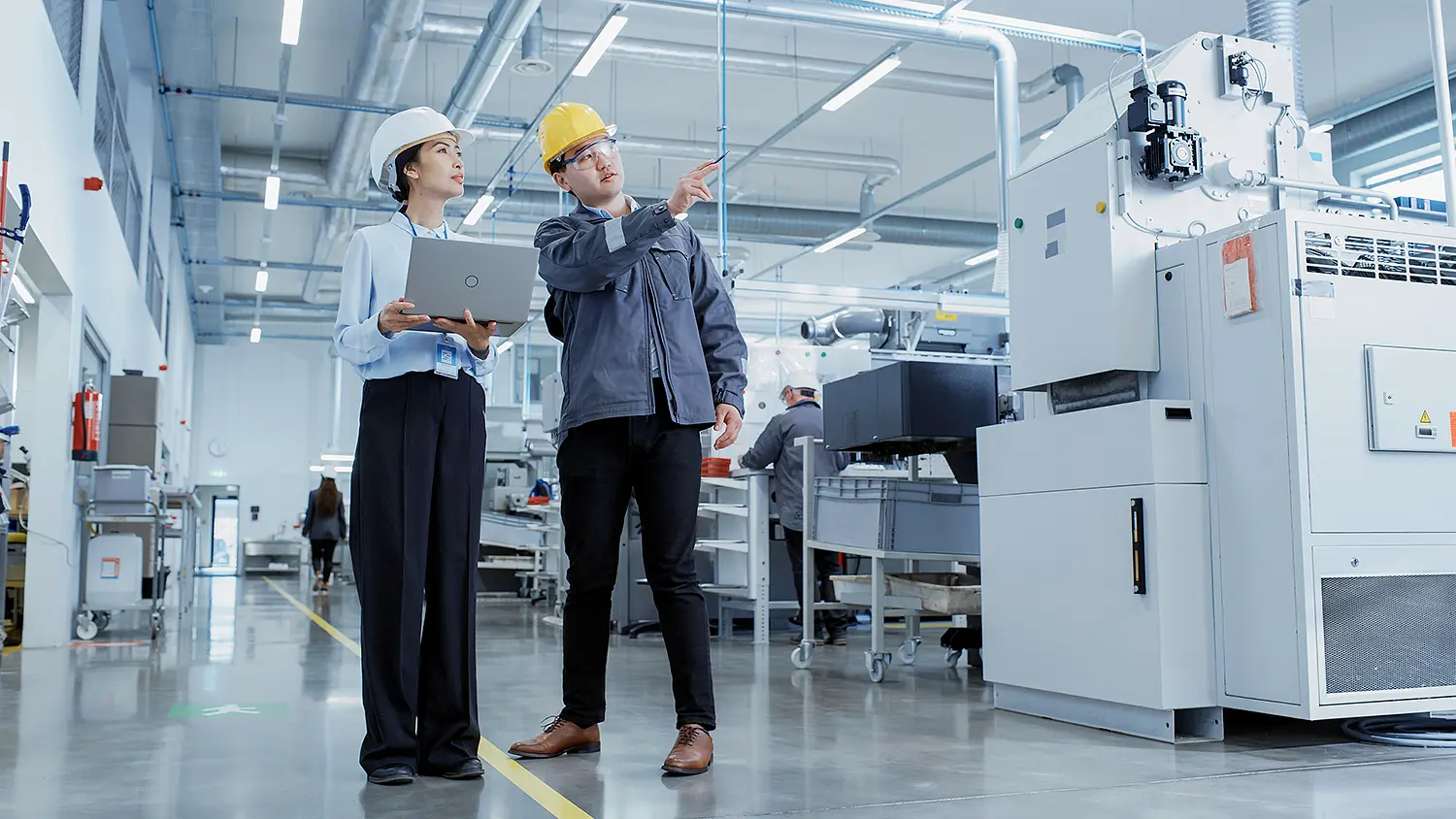 Zwei Personen stehen in einer Werkhalle. Die eine Person erklärt der anderen etwas und zeigt nach links